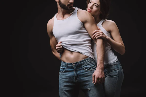 Cropped view of girl hugging and undressing man in white singlet isolated on black — Stock Photo