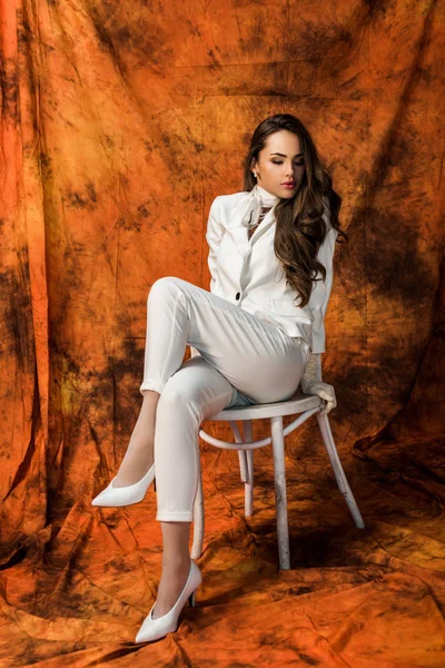 Attractive girl with long hair in white suit sitting on chair on textured background — Stock Photo