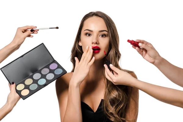 Vista recortada de las mujeres sosteniendo lápiz labial líquido, cepillo cosmético y paletas con sombras de ojos y chica sorprendida aislada en blanco - foto de stock