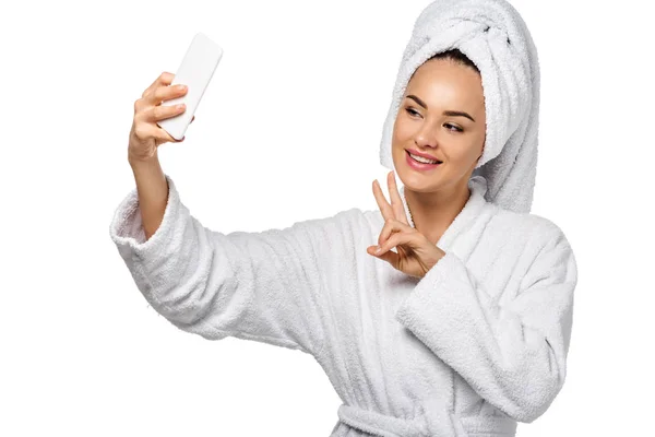 Chica atractiva en albornoz mostrando el símbolo de la paz y tomar selfie aislado en blanco - foto de stock