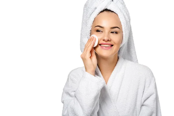 Attraktives Mädchen im Bademantel, das Gesicht mit Baumwollschwamm reinigt und isoliert auf Weiß lächelt — Stockfoto