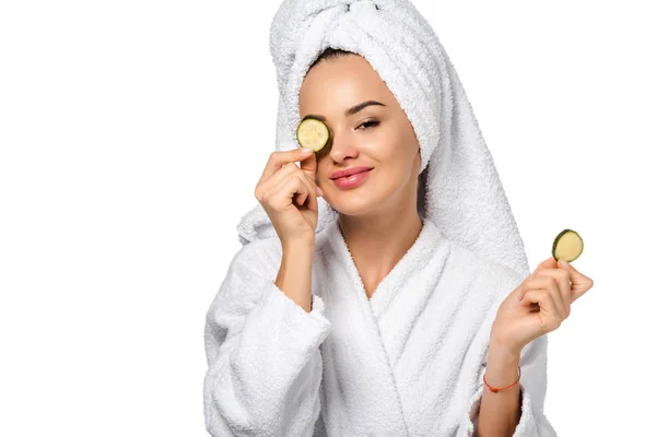 Beautiful girl in bathrobe holding cucumber slice near eye and smiling isolated on white — Stock Photo