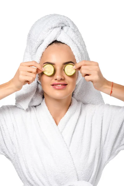 Beautiful girl in bathrobe holding cucumber slices near eyes and smiling isolated on white — Stock Photo