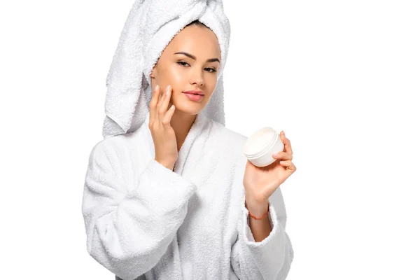 Belle fille en peignoir regardant la caméra, tenant le pot et appliquant de la crème sur le visage isolé sur blanc — Photo de stock