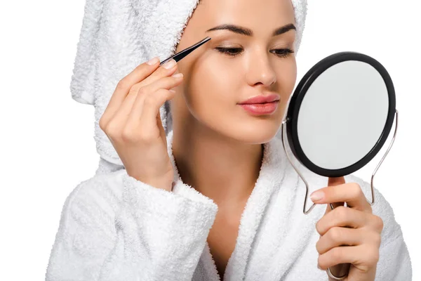 Close up view of beautiful girl in bathrobe looking at mirror and tweezing eyebrows isolated on white — Stock Photo