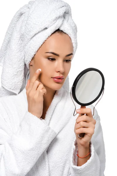 Красива дівчина в халаті дивиться на дзеркало і наносить крем на обличчя ізольовано на білому — стокове фото