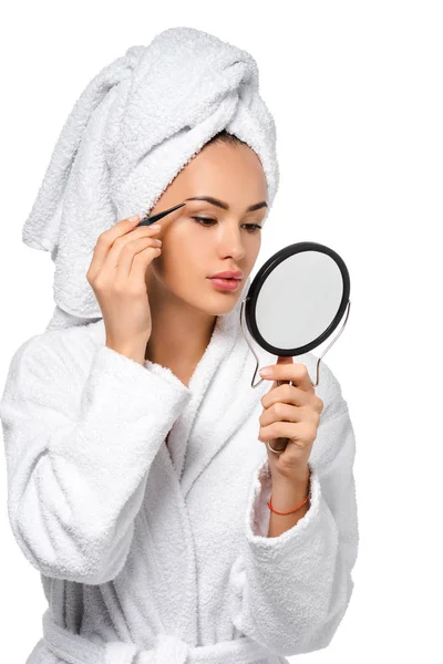 Menina bonita em roupão de banho olhando para espelho e pinça sobrancelhas isoladas no branco — Fotografia de Stock