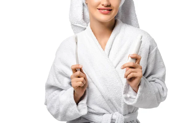Abgeschnittene Ansicht eines Mädchens im Bademantel, das Zahnbürste und Zahnpasta in der Hand hält und isoliert auf weißem Grund lächelt — Stockfoto