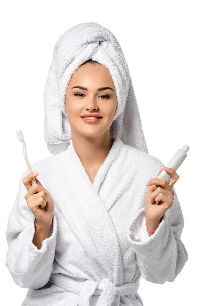 Hermosa chica en albornoz sosteniendo cepillo de dientes y pasta de dientes, sonriendo y mirando a la cámara aislada en blanco - foto de stock
