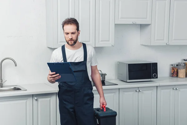 Reparador barbudo serio en el trabajo en general la celebración de la caja de herramientas y mirando el portapapeles en la cocina en casa - foto de stock