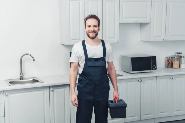 Happy repairman in working overall holding toolbox in kitchen at home — Stock Photo