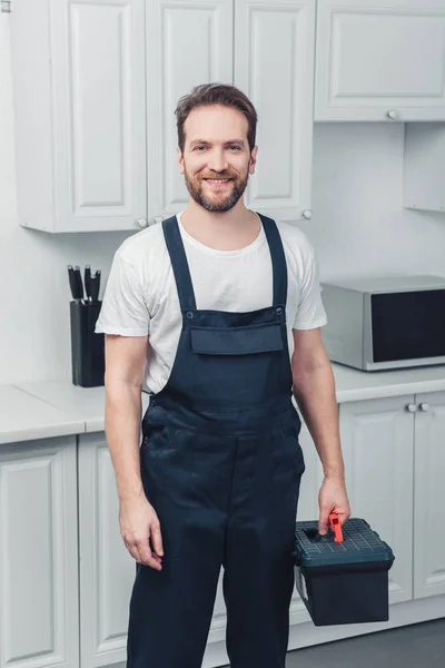 Adulto sorrindo barbudo reparador no trabalho geral segurando caixa de ferramentas na cozinha em casa — Fotografia de Stock