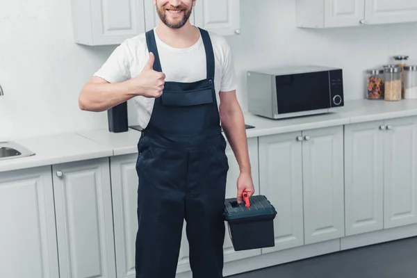 Partial view of bearded repairman in working overall holding toolbox and showing thumb up in kitchen at home — Stock Photo