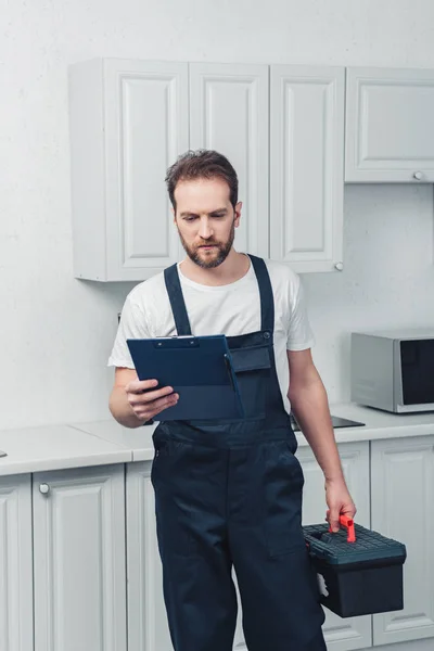 Réparateur barbu concentré dans la boîte à outils de tenue globale de travail et regardant le presse-papiers dans la cuisine à la maison — Photo de stock