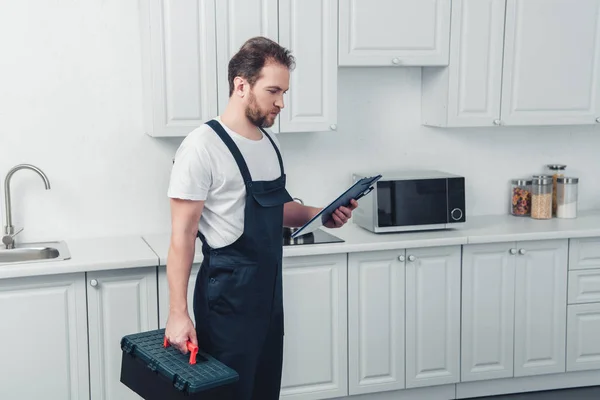 Vue latérale du réparateur adulte dans la boîte à outils de tenue globale de travail et regardant le presse-papiers dans la cuisine à la maison — Photo de stock