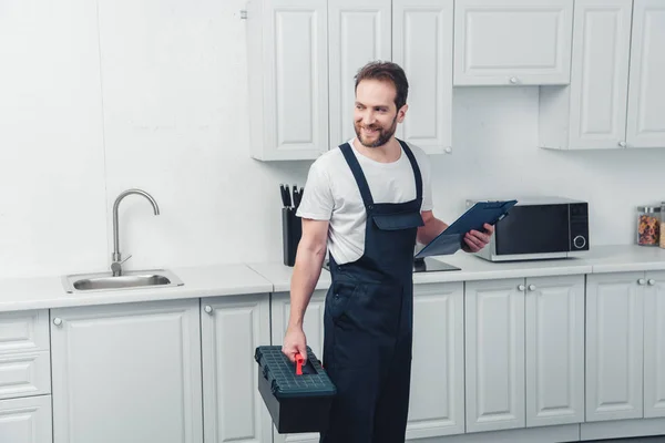 Fröhlicher erwachsener Reparateur im Arbeitsalltag mit Werkzeugkiste und Klemmbrett in der heimischen Küche — Stockfoto