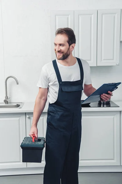 Reparador adulto feliz en el trabajo en general la celebración de caja de herramientas y portapapeles en la cocina en casa - foto de stock