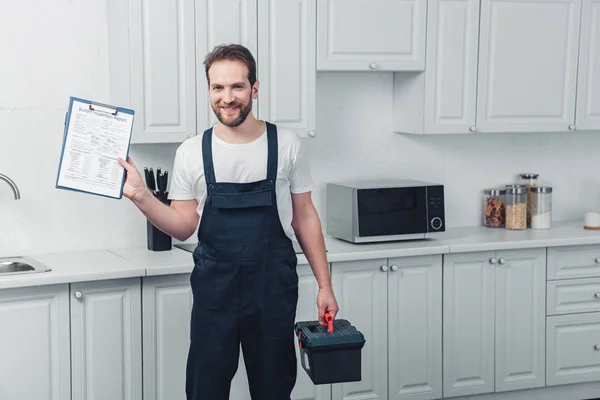 Smiling bearded repairman in working overall showing clipboard in kitchen at home — Stock Photo