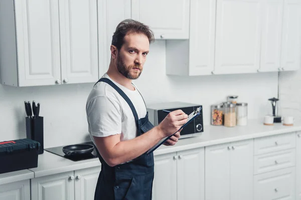 Guapo adulto barbudo reparador con portapapeles en la cocina - foto de stock