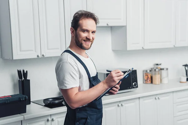 Heureux réparateur barbu adulte prenant des notes dans le presse-papiers dans la cuisine — Photo de stock