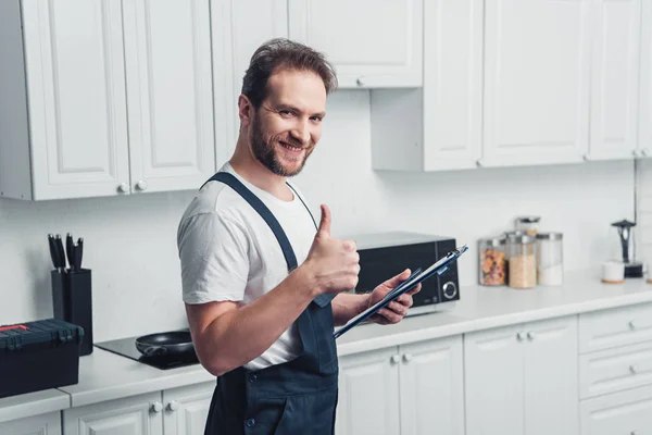 Sourire beau réparateur barbu adulte tenant presse-papiers et faisant geste pouce vers le haut dans la cuisine — Photo de stock