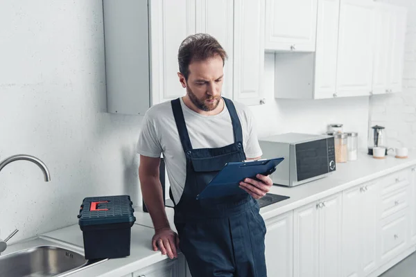Adult bearded repairman looking at clipboard in kitchen at home — Stock Photo
