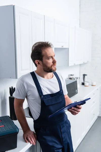 Задумчивый взрослый бородатый ремонтник с буфетом на кухне дома — стоковое фото