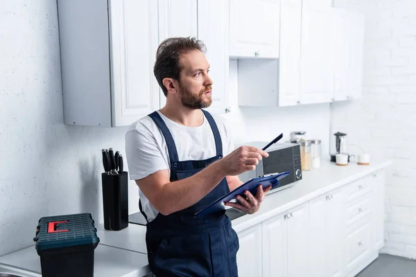 Male bearded craftsman pointing by pen and holding clipboard in kitchen — Stock Photo