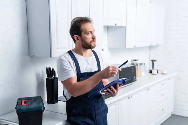 Hombre barbudo artesano señalando por pluma y sujetando portapapeles en la cocina - foto de stock