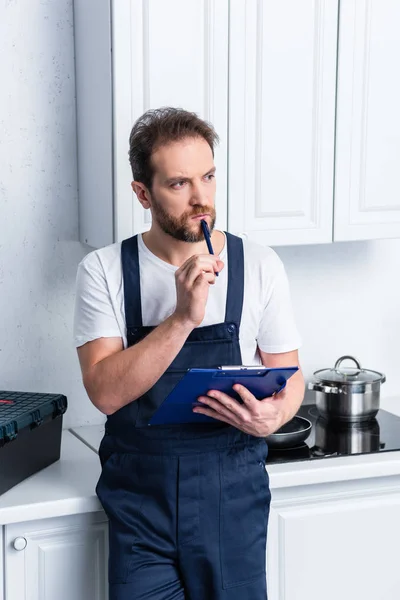 Nachdenklicher bärtiger Handwerker mit Klemmbrett steht in Küche — Stockfoto