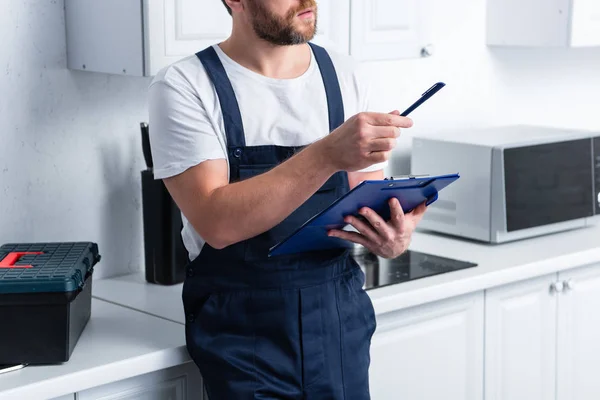 Vista parcial del artesano barbudo apuntando con la pluma y sujetando el portapapeles en la cocina - foto de stock