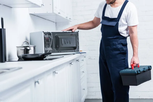 Cropped shot of handyman with toolbox checking microwave oven in kitchen — Stock Photo