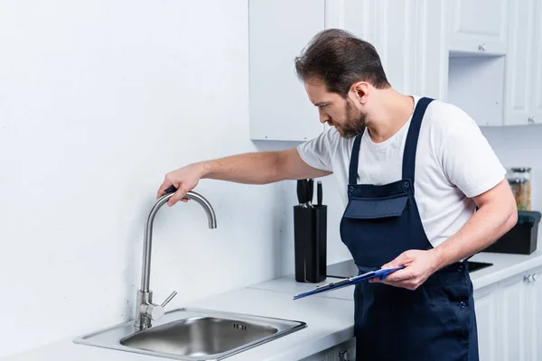 Reparador no trabalho global segurando prancheta e verificação de pia na cozinha — Fotografia de Stock