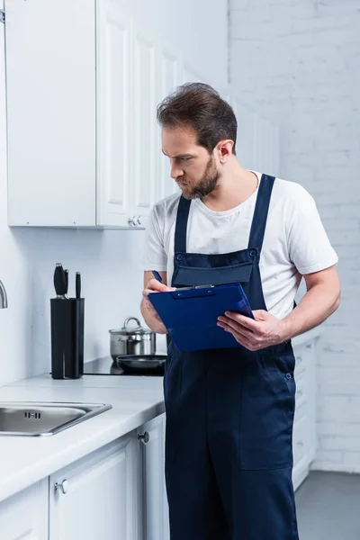 Serious adult repairman in working overall writing in clipboard and checking sink in kitchen — Stock Photo