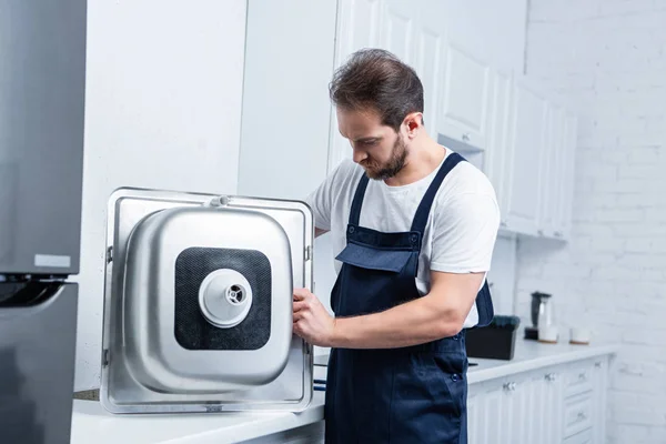 Selective focus of bearded repairman in working overall fixing sink in kitchen — Stock Photo