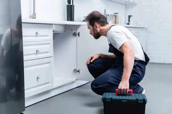 Side view of adult bearded repairman with toolbox checking sink in kitchen — Stock Photo