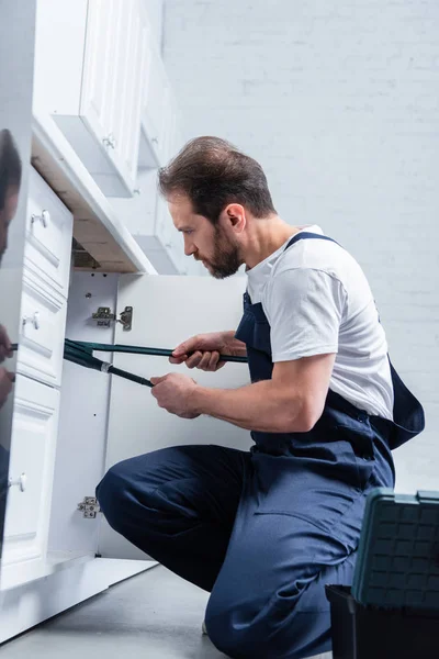 Focused repairman in working overall fixing sink with pair of nippers in kitchen — Stock Photo