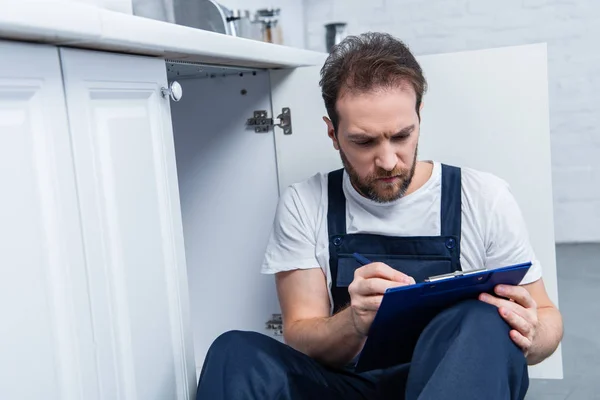 Serious adult craftsman writing in clipboard while sitting on floor in kitchen — Stock Photo