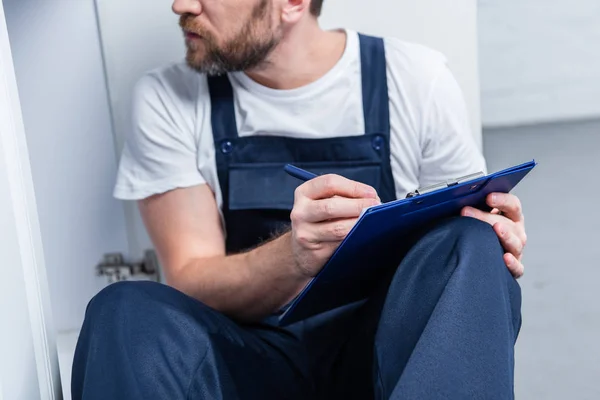 Teilansicht eines erwachsenen Handwerkers, der in Klemmbrett schreibt, während er in der Küche auf dem Boden sitzt — Stockfoto