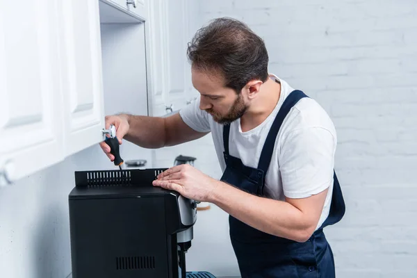 Bearded handyman in working overall repairing coffee machine by screwdriver in kitchen — Stock Photo