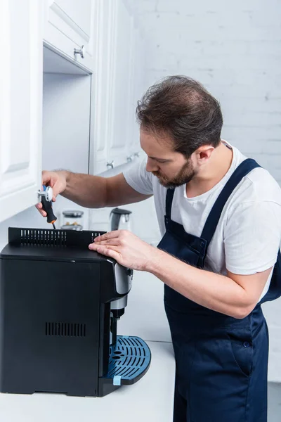 Adult handyman in working overall repairing coffee machine by screwdriver in kitchen — Stock Photo