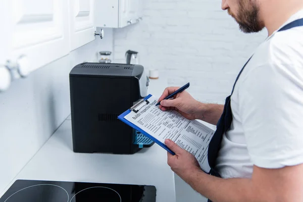 Teilansicht eines Handwerkers, der in der Nähe einer kaputten Kaffeemaschine in der Küche Notizen in Klemmbrett macht — Stockfoto