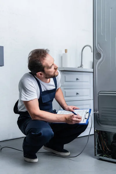 Craftsman in working overall writing in clipboard near broken refrigerator in kitchen — Stock Photo