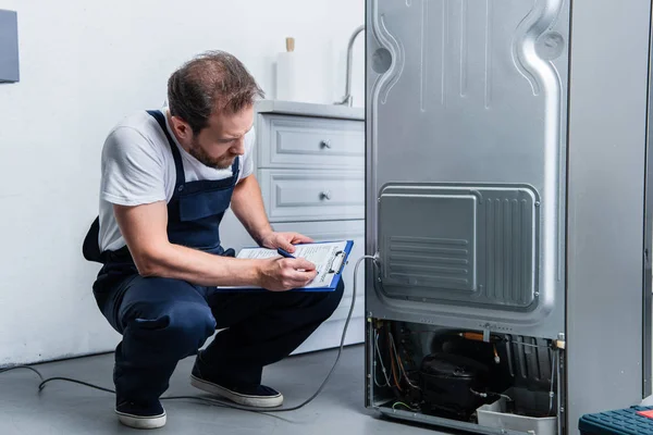 Adult craftsman in working overall writing in clipboard near broken refrigerator in kitchen — Stock Photo