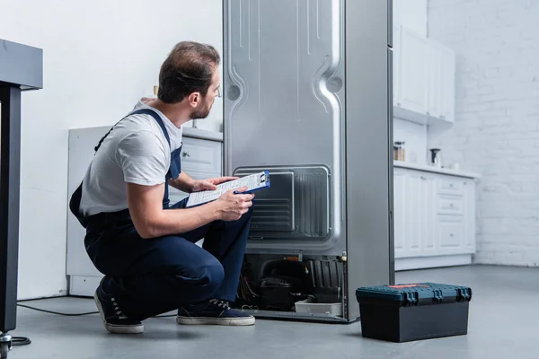 Handwerker in Arbeitskleidung schreibt in Klemmbrett neben kaputtem Kühlschrank in Küche — Stockfoto