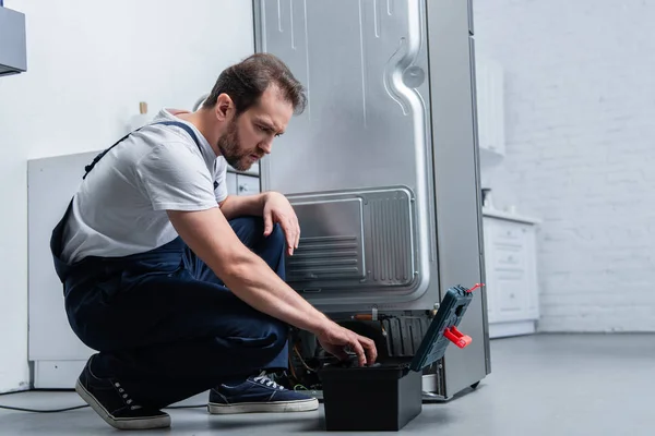 Repairman in working overall near taking tools from toolbox broken refrigerator in kitchen — Stock Photo