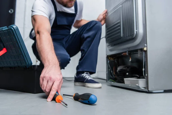 Recortado disparo de reparador en el trabajo en general tomando destornillador del piso cerca de nevera rota en la cocina - foto de stock