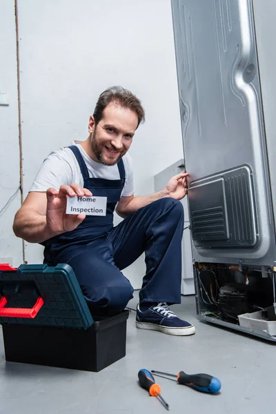 Sonriente adulto artesano mostrando tarjeta con letras de inspección en casa mientras está sentado cerca de la nevera rota - foto de stock