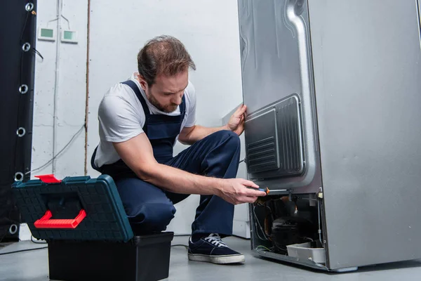 Adult bearded repairman in working overall fixing refrigerator by screwdriver in kitchen — Stock Photo