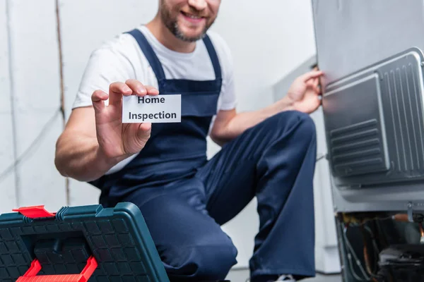Частичный вид взрослого ремесленника, показывающего карточку с надписью домашний осмотр, сидя рядом со сломанным холодильником — стоковое фото